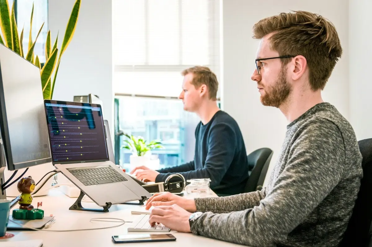 Deux hommes travaillant sur des ordinateurs avec du code à l'écran, dans un bureau lumineux