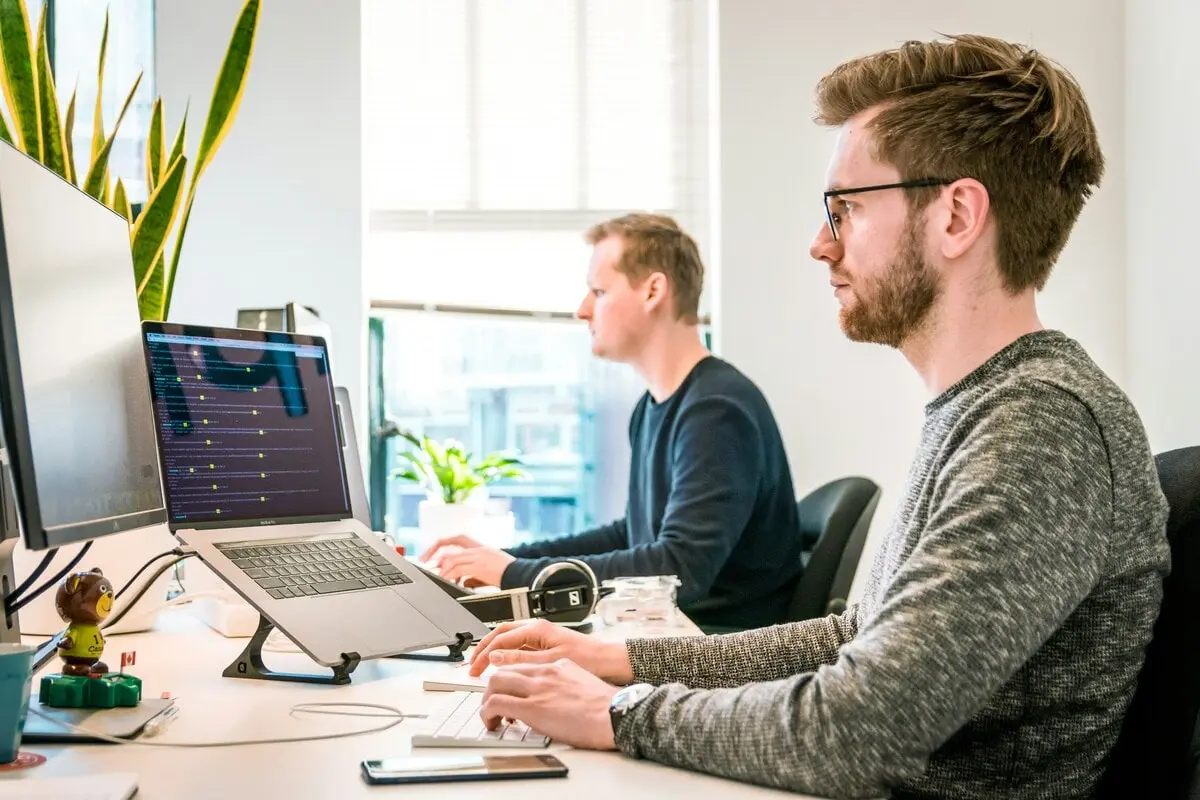 Deux hommes travaillant sur des ordinateurs avec du code à l'écran, dans un bureau lumineux